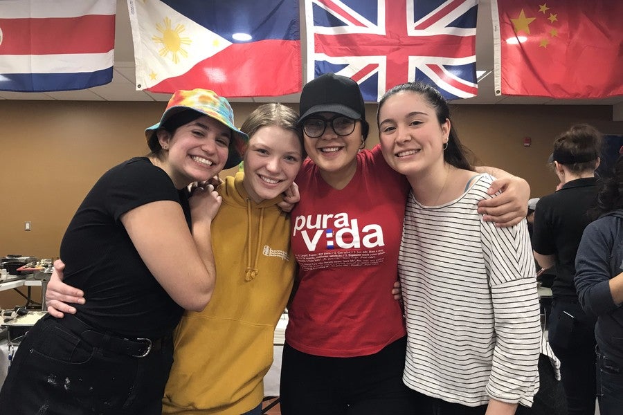 Four students smile at the camera in front of a wall of international flags.