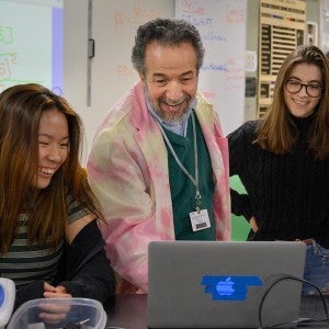 A teacher dressed in a brightly-colored coat smiles as he helps two students with their classwork.