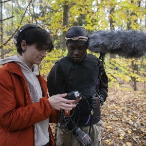 Two Film & New Media students, surrounded by forest, look at a camera's viewfinder.