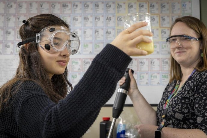 A student wearing safety goggles holds up a beaker filled with yellow liquid while another student watches.