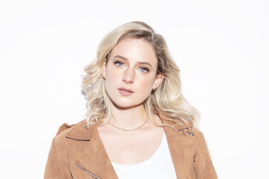 A young woman gazes at the viewer. Her hair is blonde and she wears a suede jacket with a white shirt underneath.