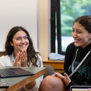 Two students smile, one clapping and the other looking at her writing on a laptop.