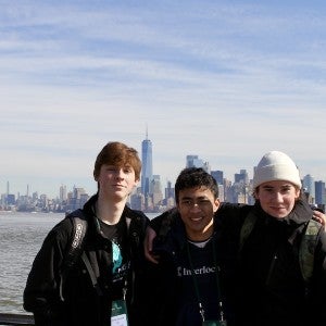 Three male students dressed in warm coats stand by the water, with the New York skyline behind them.