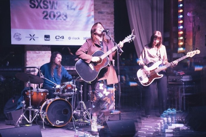 A young woman dressed in a brown jacket and flowered skirt plays guitar and sings into a microphone. She is accompanied by a guitarist and a drummer.