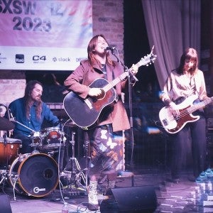 A young woman dressed in a brown jacket and flowered skirt plays guitar and sings into a microphone. She is accompanied by a guitarist and a drummer.