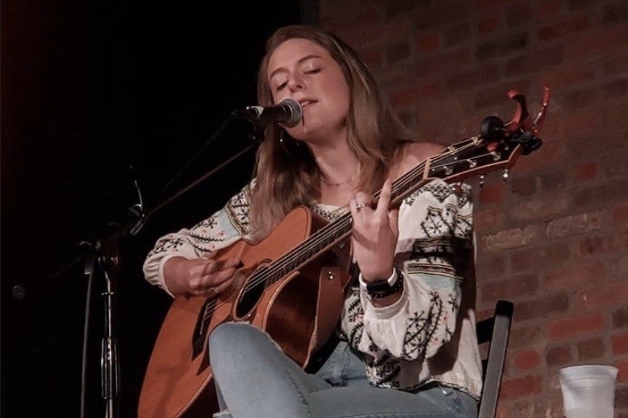 A young woman with blond hair closes her eyes, strums a guitar, and sings into a microphone.