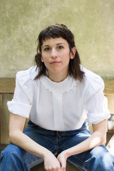 A dark-haired woman wearing a white button-down shirt and jeans gazes at the viewer.
