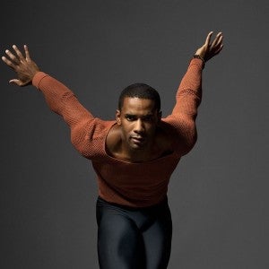 A man dressed in orange and black poses with his arms flung up behind him.