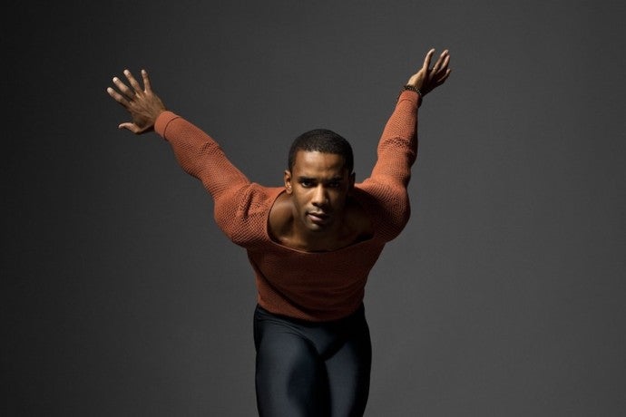 A man dressed in orange and black poses with his arms flung up behind him.