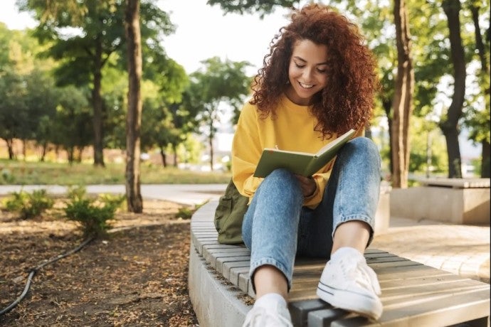 A student in a yellow sweater and jeans smiles as she reads a book.